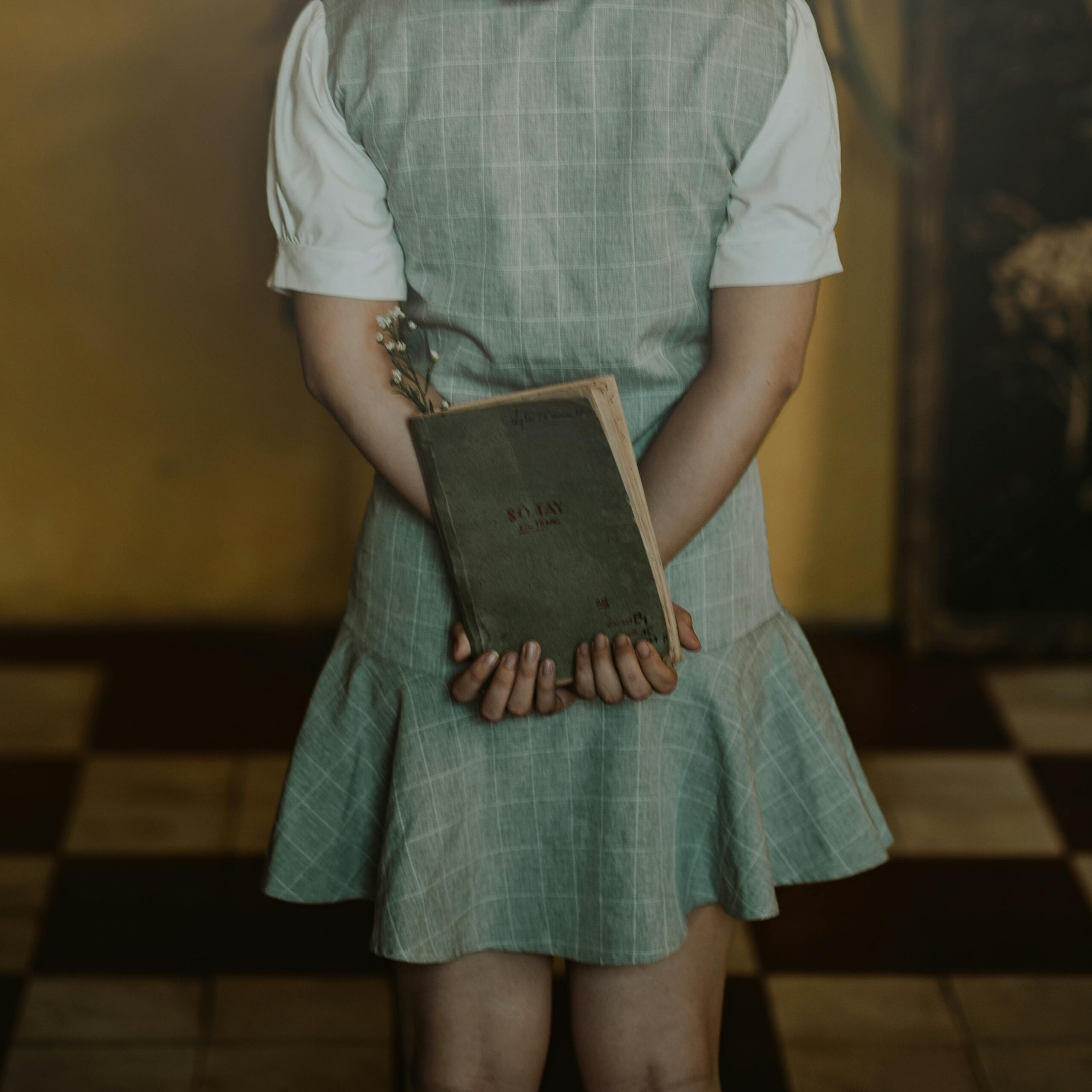 A young woman holding a book behind her back in a vintage dress, standing indoors.